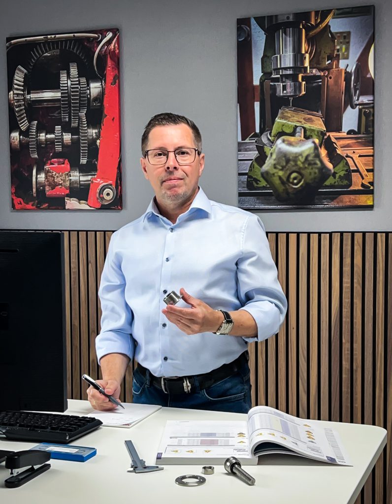 Mann am Schreibtisch hält Metallteil und Stift neben Computer, Zeichnungen und Werkzeug